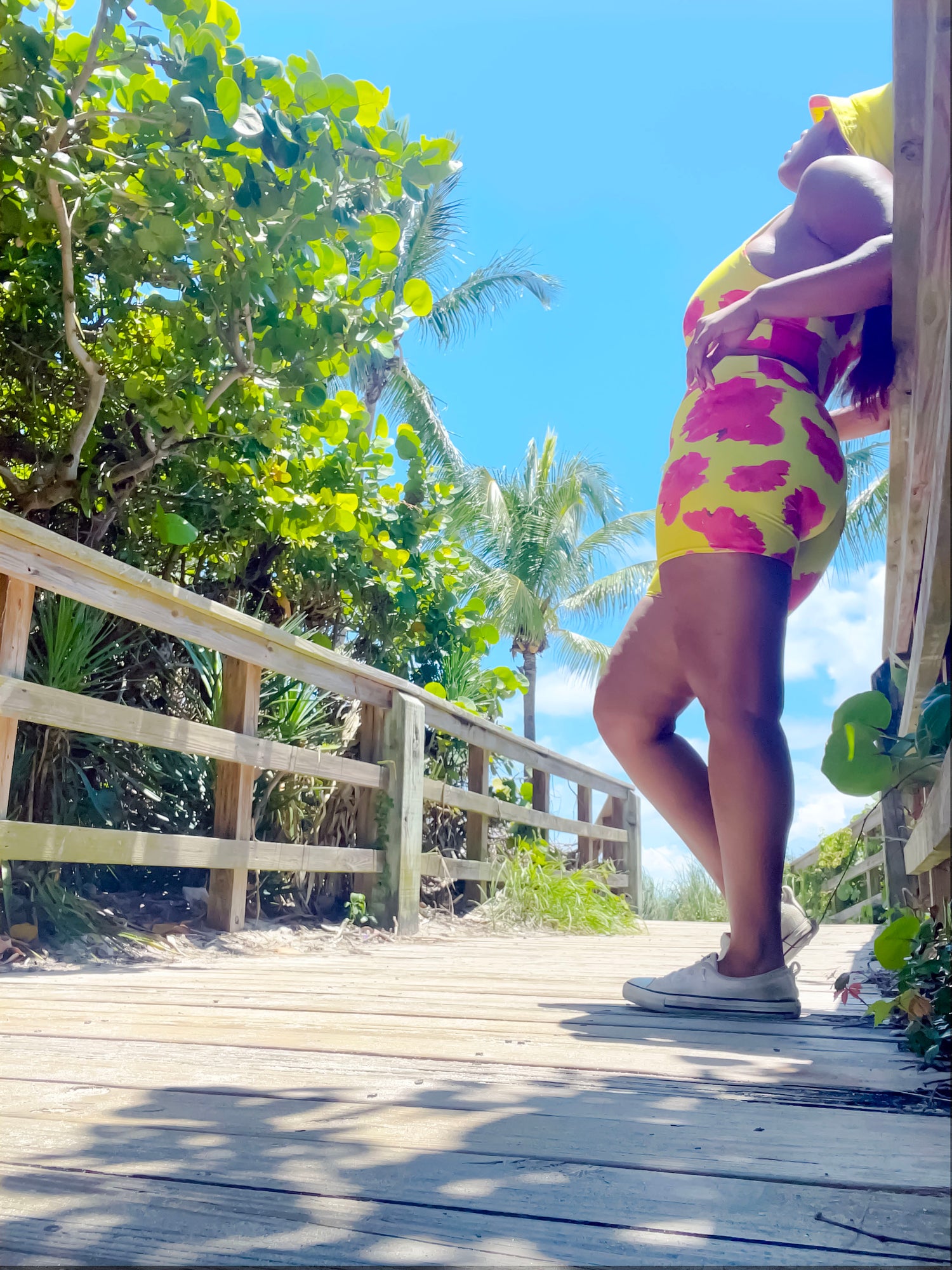 Beach boardwalk scene with girl wearing yellow biker shorts with red hibiscus flowers on them and a matching sports bra and yellow reversible bucket hat. looking at blue sky.